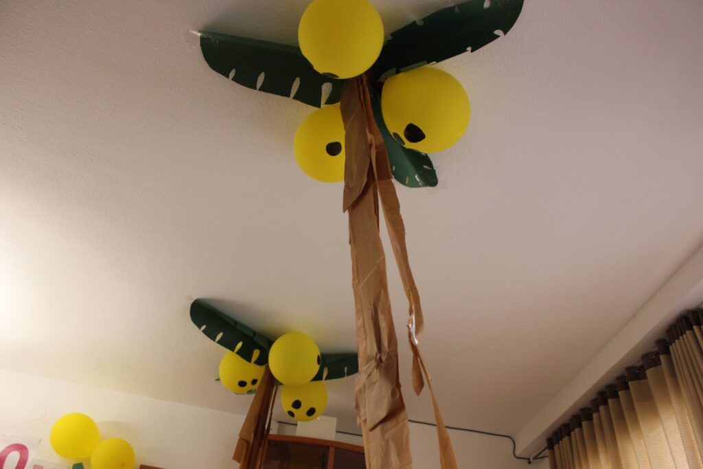fiesta de cumpleaños - Las palmeras tropicales: hojas hechas con cartulina pegadas al techo, globos amarillos con agujeros pintados con un rotulador negro y tiras marrones de papel de seda.